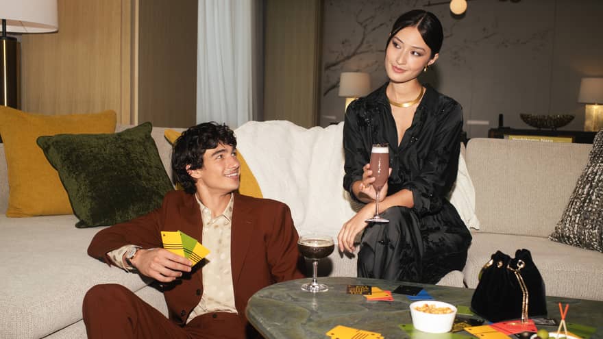 Two people sitting on a sofa with drinks, surrounded by snacks and game cards, enjoying a casual gathering in a stylish living room