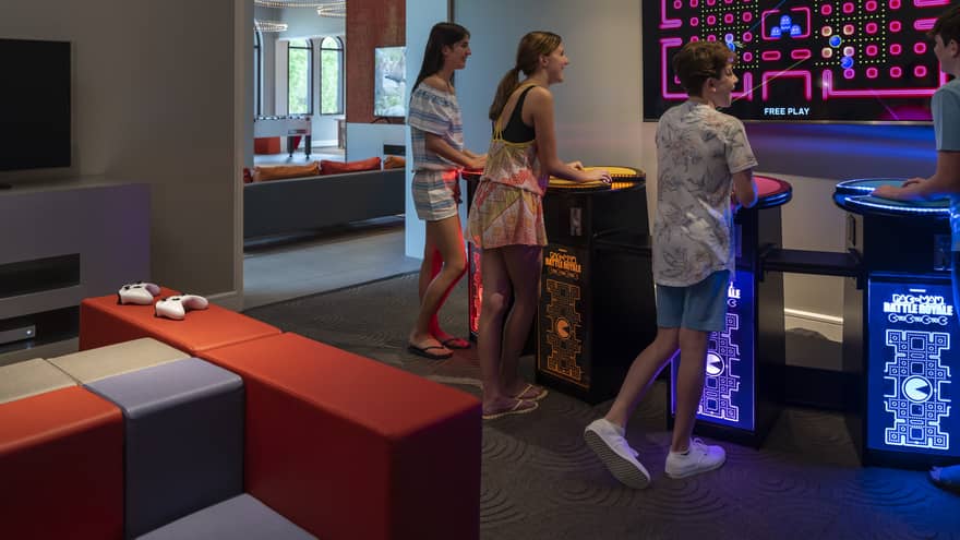 Four smiling teenagers stand at multi-coloured video-game pedestals facing a large TV screen in a dimly-lit games room.