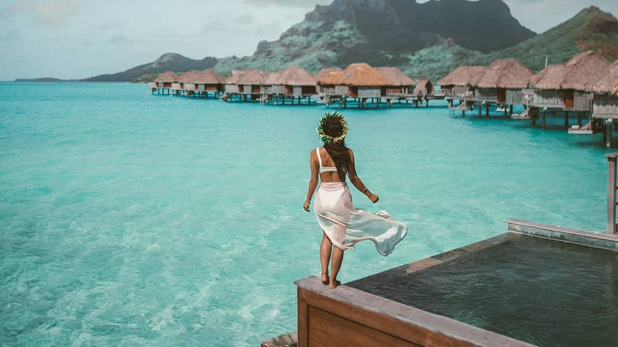 Woman stands at the edge of the infinity pool overlooking the overwater bungalows, mountains in distance