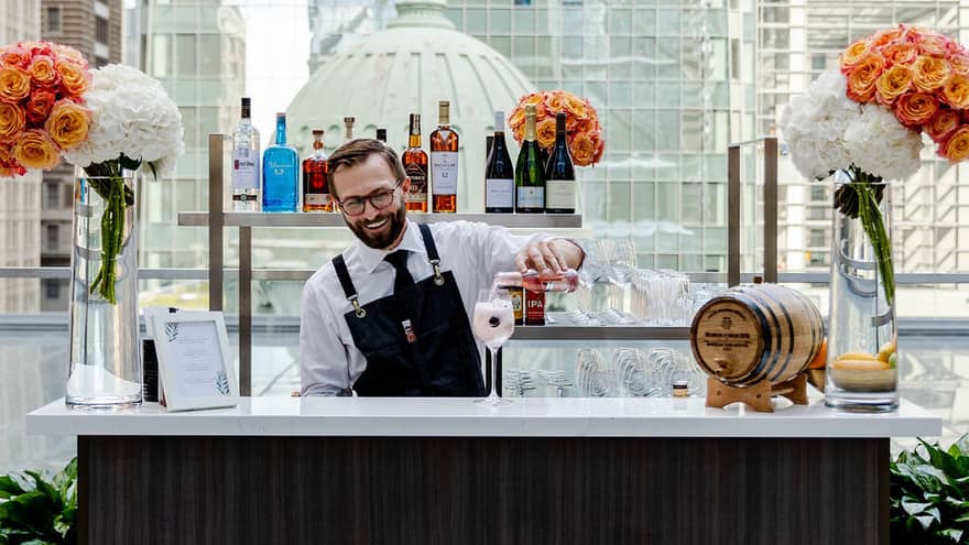 A bartender with orange flowers near him in a black apron making a pink cocktail near a large window