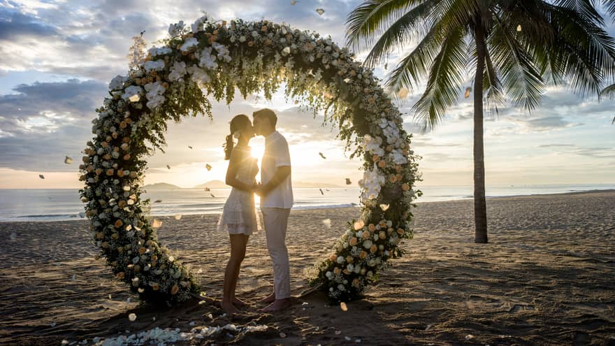 Bride and groom kiss under round wedding altar with flowers on beach at sunset