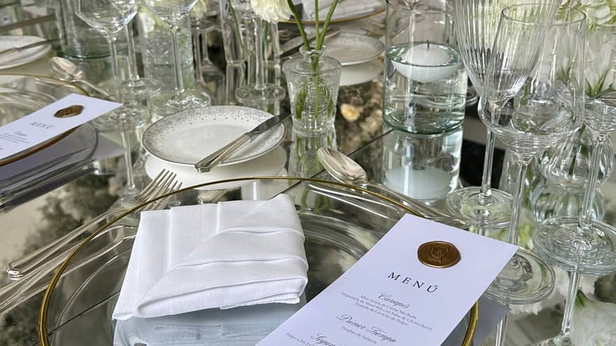 A close-up of a glass table set with clear dishes and stemware, flatware, intricately folded white napkins, white floral decor, and candles, with an elegant printed menu set on each plate.