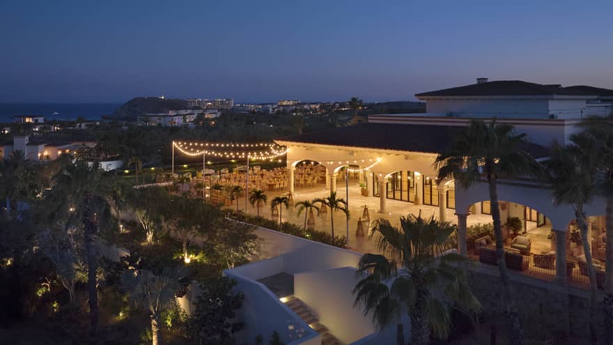 A an outdoor event space brightly lit in the evening, set among palm trees and next to a building