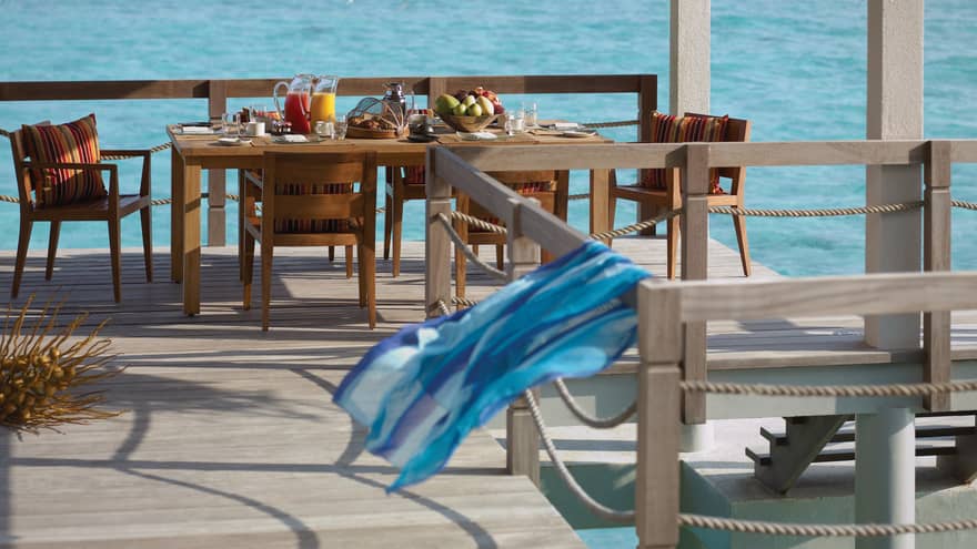 Long wood patio dining table with fresh juice, fruit, pastries, blue scarf blowing in breeze