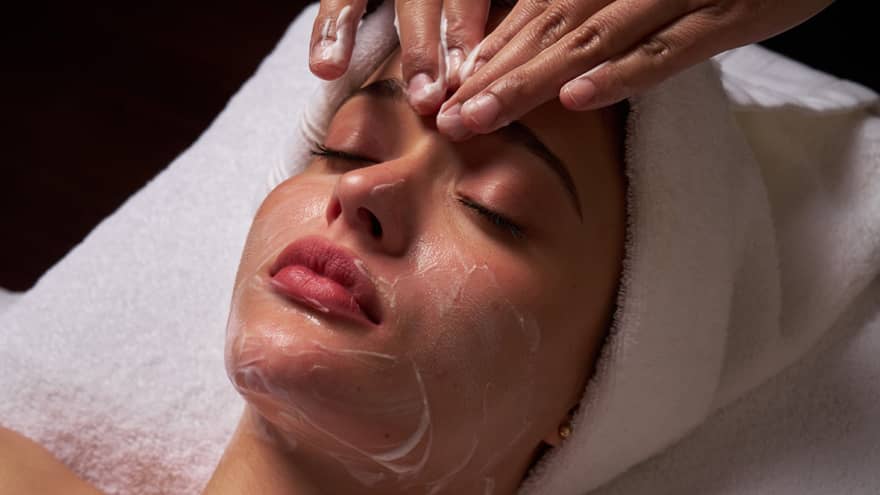 Person lies on massage table covered in a white cloth from shoulders down with hair wrapped in white towel while a pair of hands massages their forehead