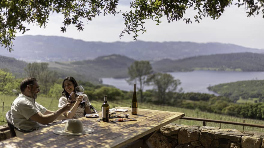 A couple smile and clink wine glasses as they sit at a rustic outdoor table overlooking a green valley and hill-lined lake.