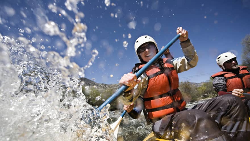 Smiling woman wearing red life vest holds paddles as waves crash over raft