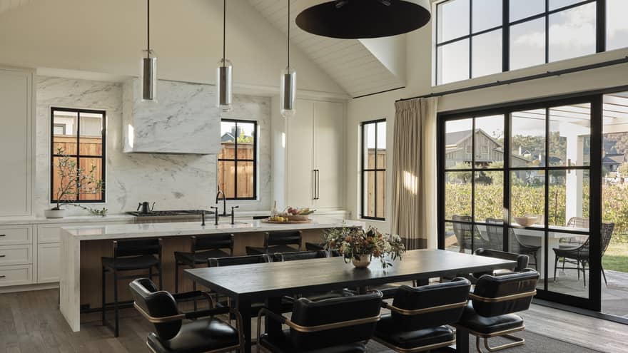Open kitchen and dining area with marble island, dark wood table, black leather chairs, pendant lights, windows and sliding doors leading to outdoor patio