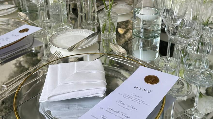 A close-up of a glass table set with clear dishes and stemware, flatware, intricately folded white napkins, white floral decor, and candles, with an elegant printed menu set on each plate.