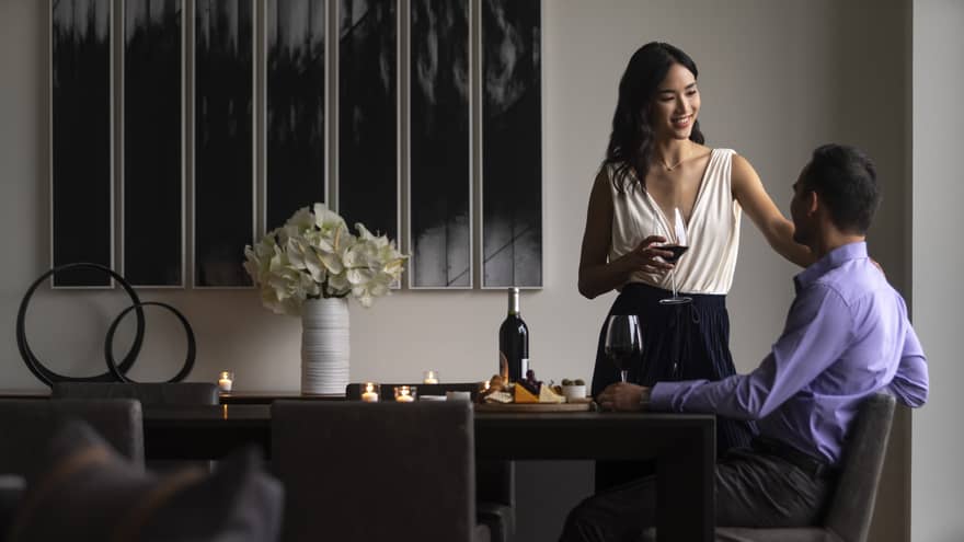 A couple drinking wine and talking in their hotel room, the man is sitting while the woman has her hand on his shoulder lovingly.