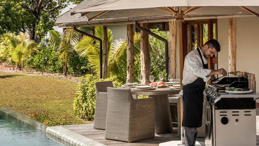 Chef wearing apron cooks at barbecue on deck by dining table, pool