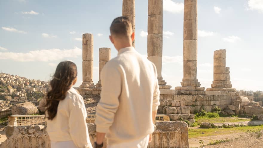 Two people both dressed in white pants and sweaters stand looking at ancient ruins