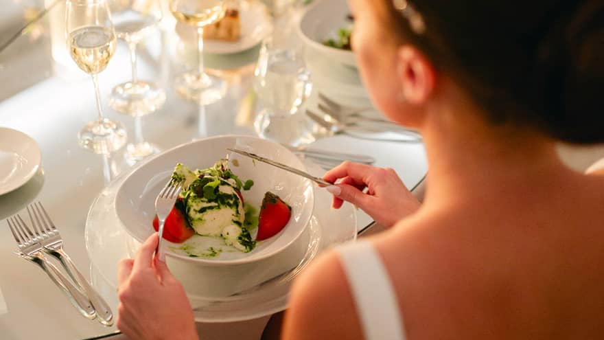 Person in bridal gown sits at round table set for dinner, fork and knife posed to dig into a meal