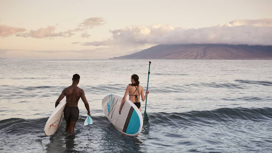 Two people standing in the ocean holding paddleboards with mountains in the background