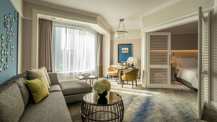 Four Seasons Executive Suite L-shaped sofa by sunny window with white curtains, table, wood shutters