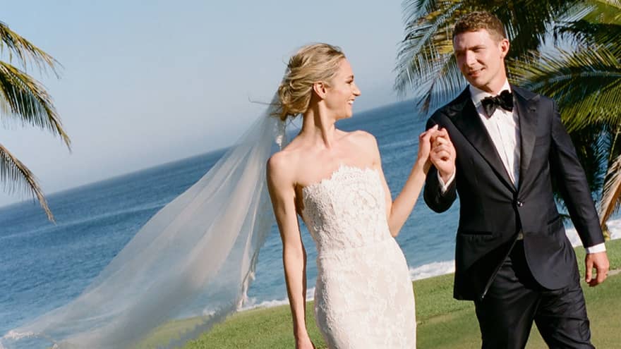 A bride in a white dress and veil and a groom in a tuxedo walk across a verdant lawn.