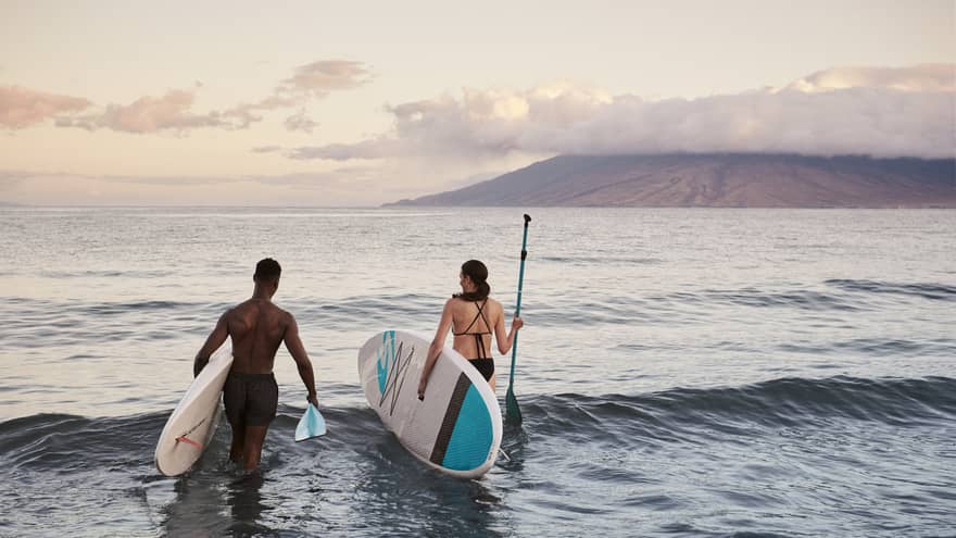 Two people standing in the ocean holding paddleboards with mountains in the background