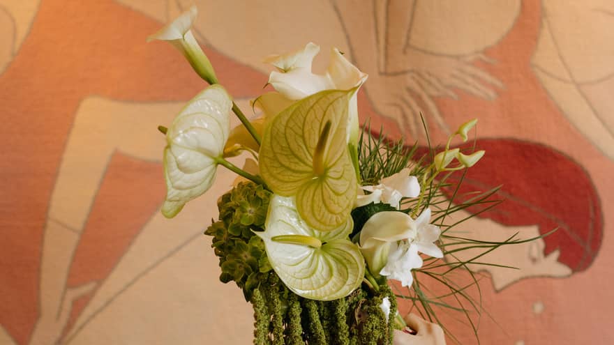 An extended arm hold a wedding bouquet made with white orchids and greenery in front of a pink-and-white mural