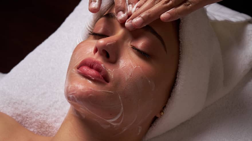 Person lies on massage table covered in a white cloth from shoulders down with hair wrapped in white towel while a pair of hands massages their forehead