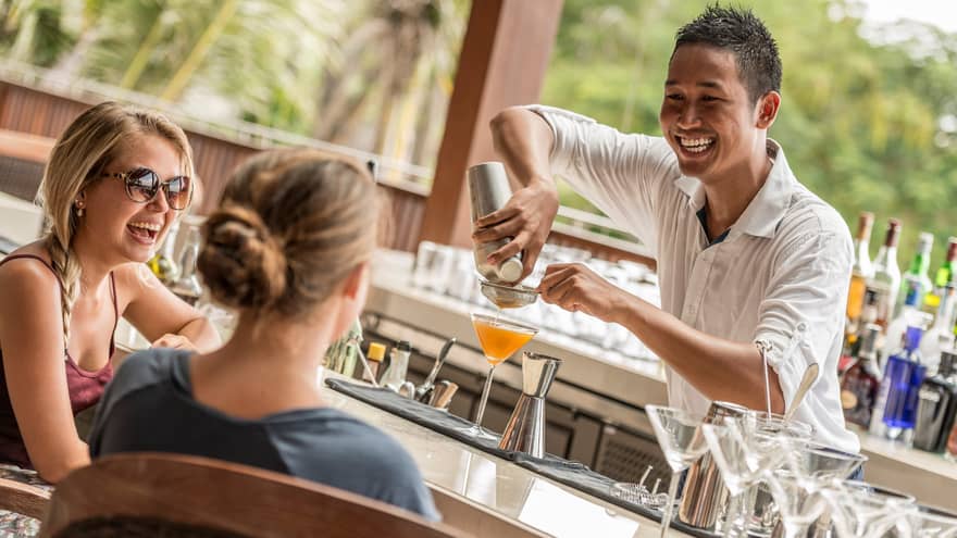Smiling mixologist pours orange cocktail in martini class by two women at bar