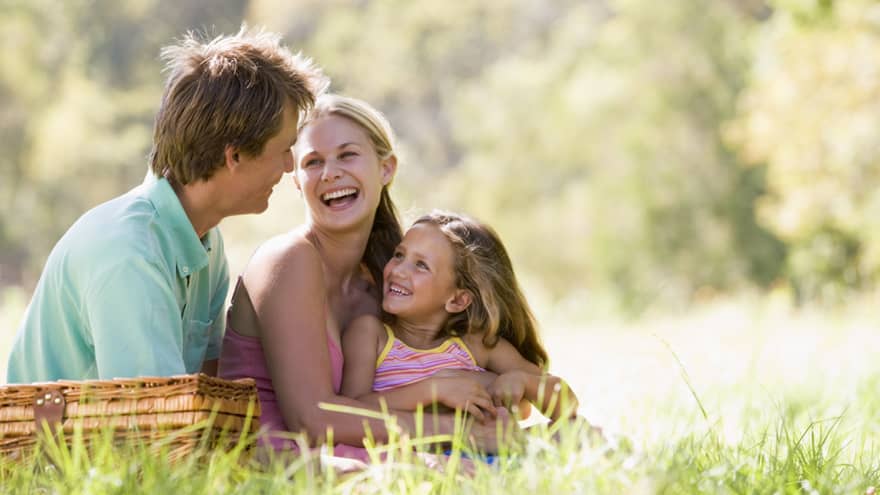 Smiling man, woman and young girl sit in tall grass by picnic basket on sunny day
