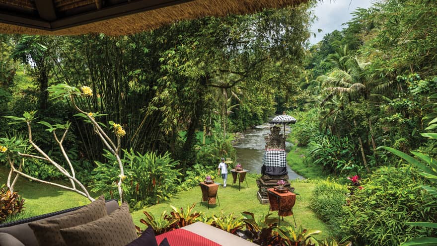 Server walks up lawn from river carrying tray of Champagne toward villa patio with chair, pillows