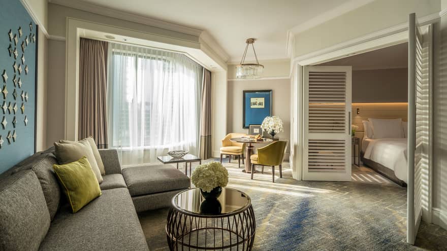 Four Seasons Executive Suite L-shaped sofa by sunny window with white curtains, table, wood shutters
