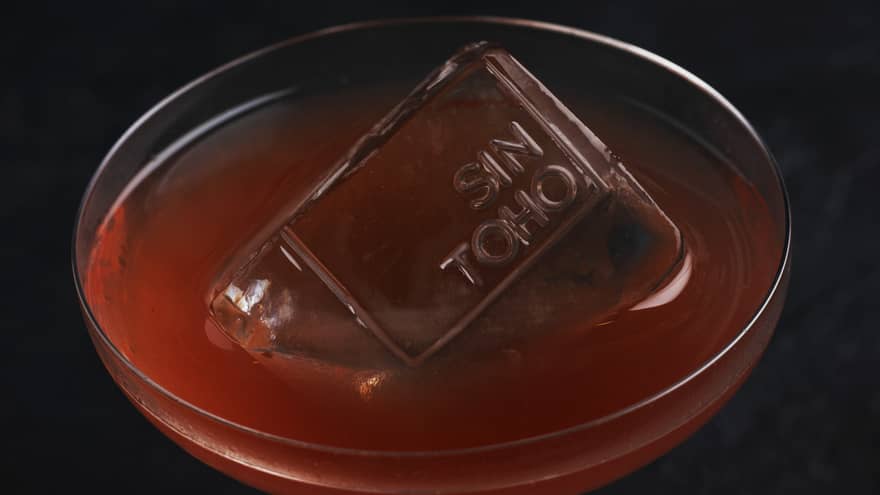 Orange-coloured beverage in a glass with a Sintoho branded ice cube.