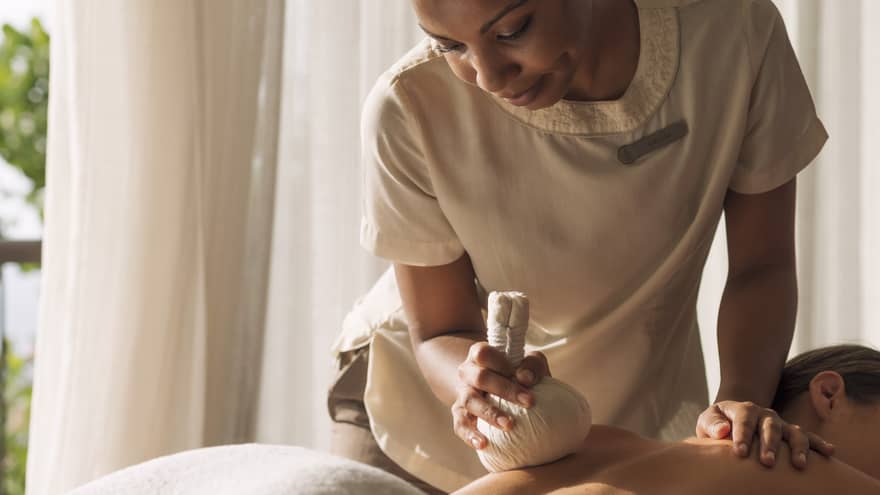 Woman holds stuffed round cloth over woman's bare shoulders as she lies on massage table