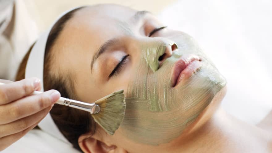 Spa staff holding brush paints green mask on woman's cheeks, chin, nose 
