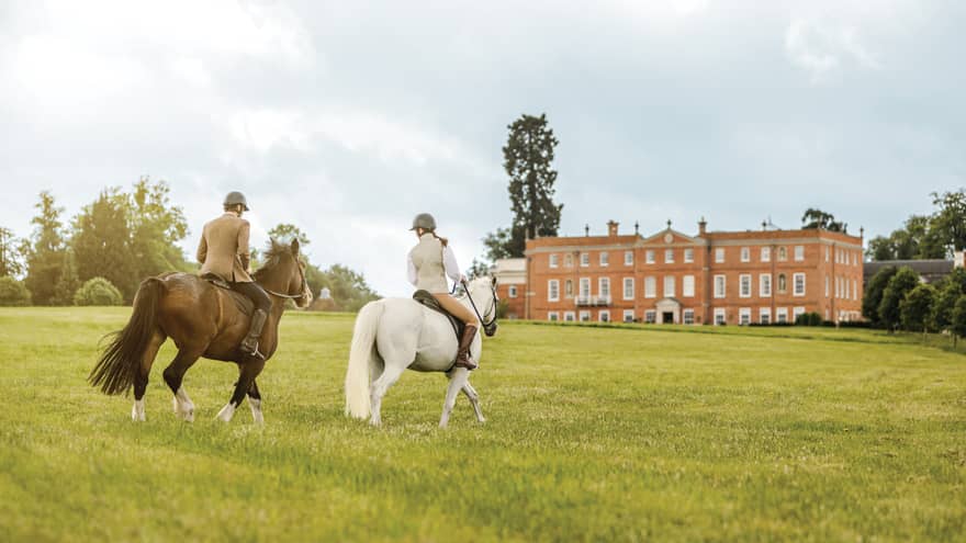 Couple rides horses across green English field to Four Seasons Hotel Hampshire building