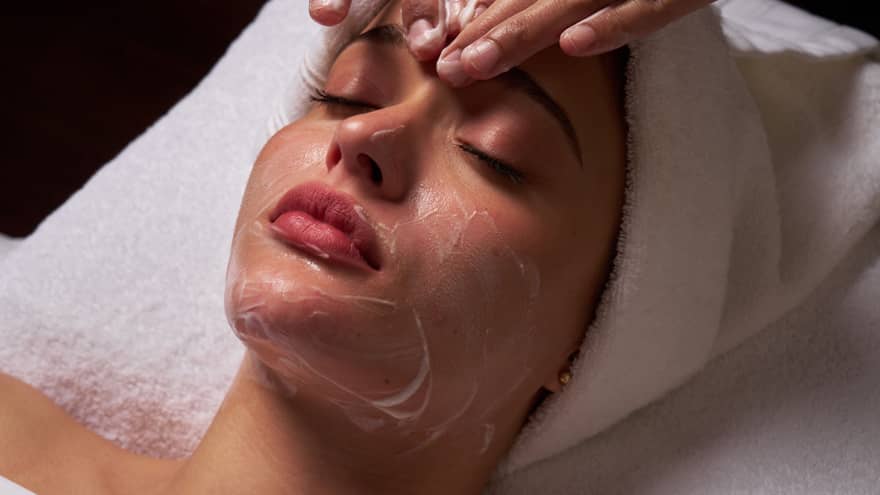Person lies on massage table covered in a white cloth from shoulders down with hair wrapped in white towel while a pair of hands massages their forehead