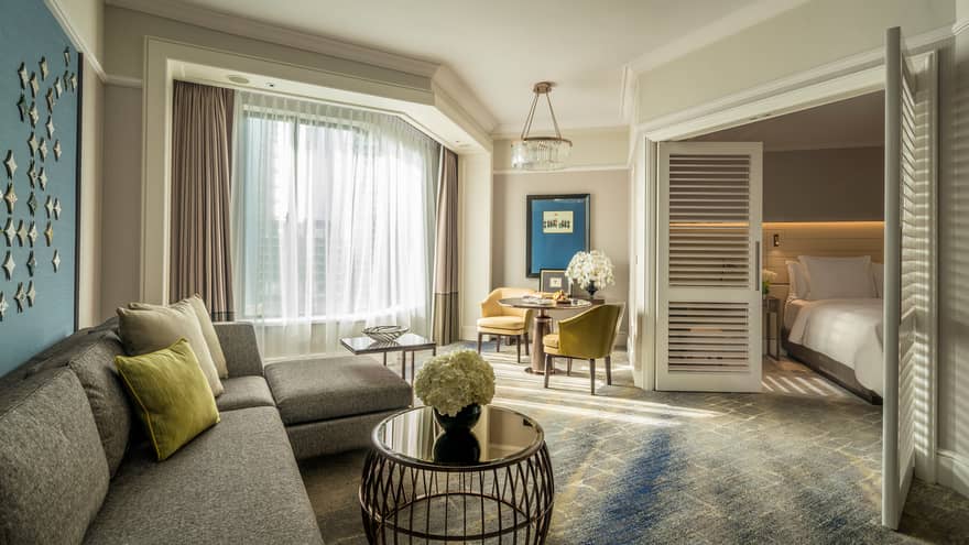 Four Seasons Executive Suite L-shaped sofa by sunny window with white curtains, table, wood shutters
