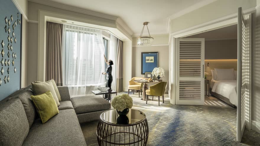 Hotel suite with a living area, separate bedroom, and large windows. A staff member adjusts the curtains, creating a bright and inviting atmosphere.