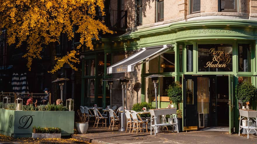 A corner cafe building made of stone and green painted metal, there are trees with orange leaves and chairs outside.