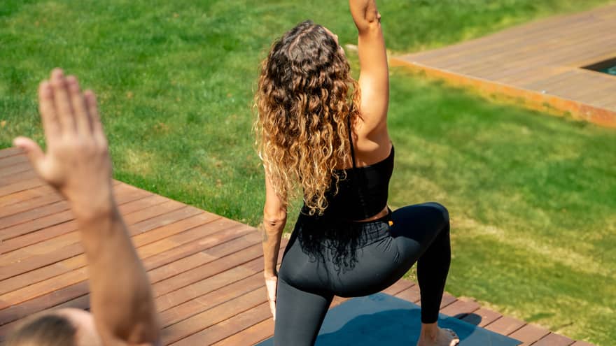 Two people in yoga pose practise on outdoor wooden deck overlooking lush green grass