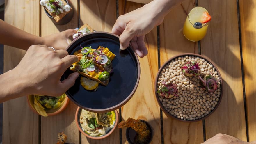 Four arms reach over a table filled with small dishes, each holding or passing along a different food item