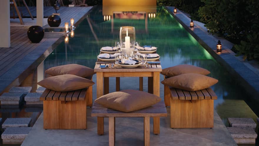 Small wood patio table with brown cushions on stool, candle in large votive, pool in background
