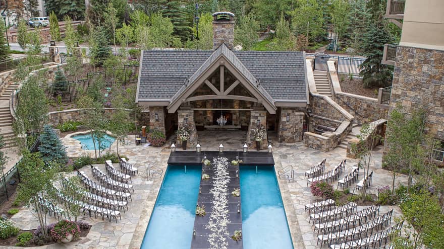 Outdoor wedding setup with a long black aisle over a pool, lined with flower petals, leading to a stone chapel. Surrounded by trees and mountains. 