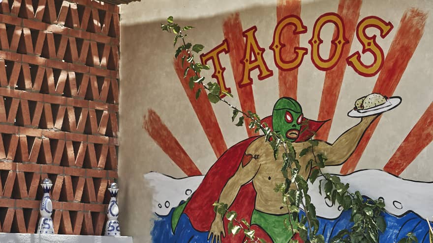 A painting of a green wrestler holding a taco on a plate behind a round chair outside.