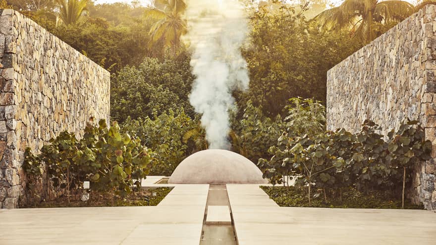 White steam rises from a small cement dome nestled between two large stone walls and surrounded by dense dark-green foliage.