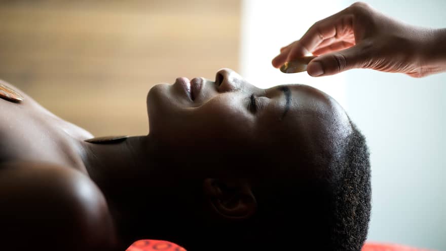 Hand placing stone on woman's forehead as she smiles, face-up on spa table
