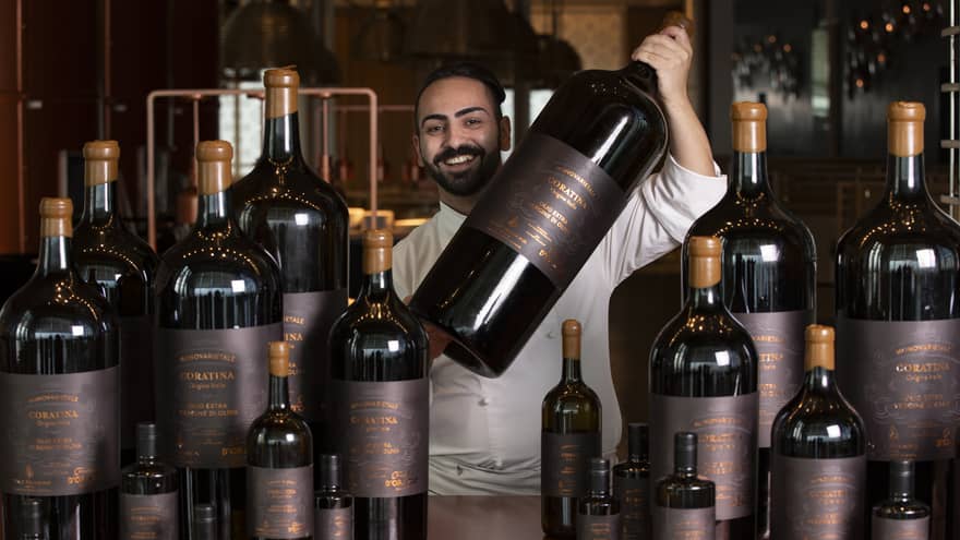 A chef smiles as he holds up a giant bottle of olive oil next to a table filled with bottles of varying sizes