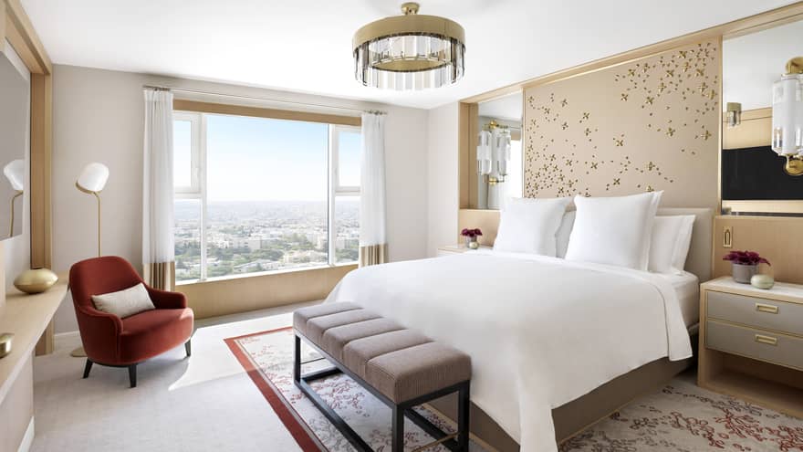 A large floral-themed bedroom with a bed in the centre, and a burgundy chaise beside a large bay window overlooking the city. 