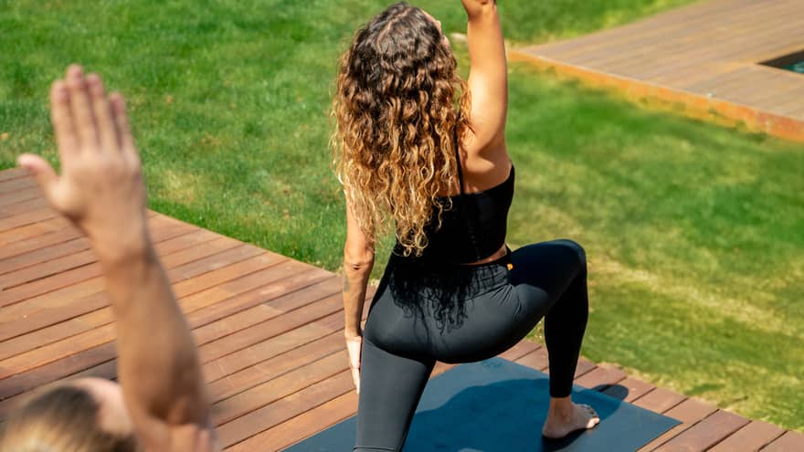 Two people in yoga pose practise on outdoor wooden deck overlooking lush green grass