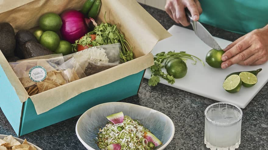 A person in a teal apron cutting lemon, there is a box of fruit and vegetables on the counter next to them.