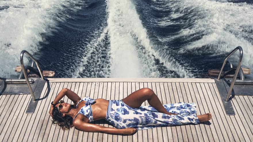 Woman sunbathes on the deck of a boat, the frothy and foaming wake visible for miles
