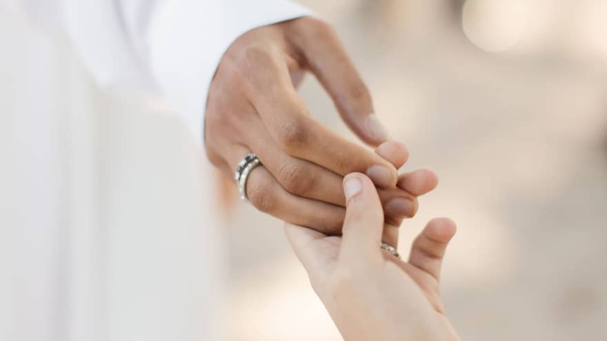 Two hands reaching out to each other and gently touching fingers, each wearing a wedding band