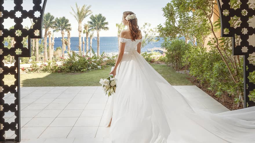 Bride in flowing white gown stands beneath outdoor carved arch