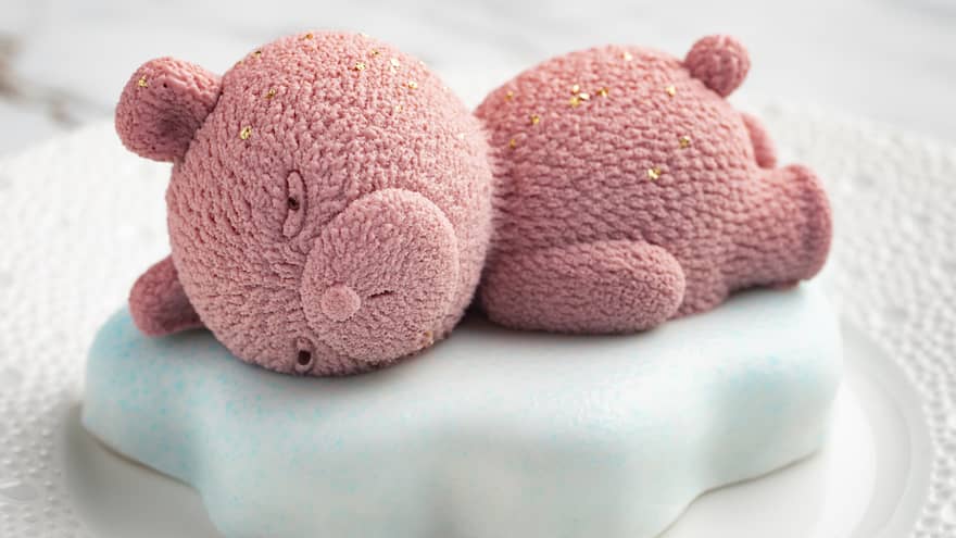 Pink teddy bear made out of chocolate mousse lays on top of a white-and-blue frosted cloud of lemon spounge cake